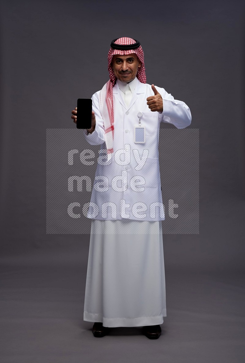 Saudi man wearing thob with lab coat and shomag with pocket employee badge standing showing phone to camera on gray background