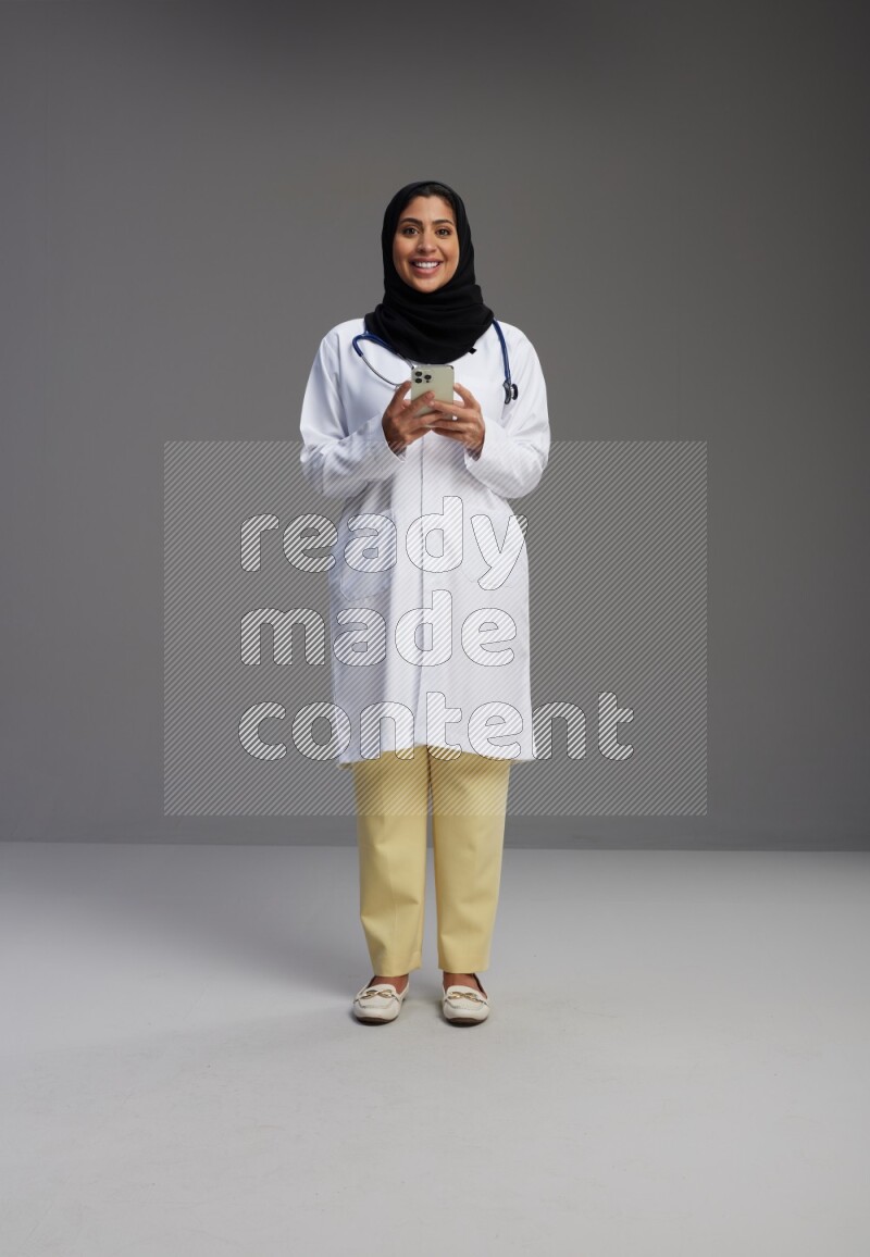 Saudi woman wearing lab coat with stethoscope standing texting on phone on Gray background