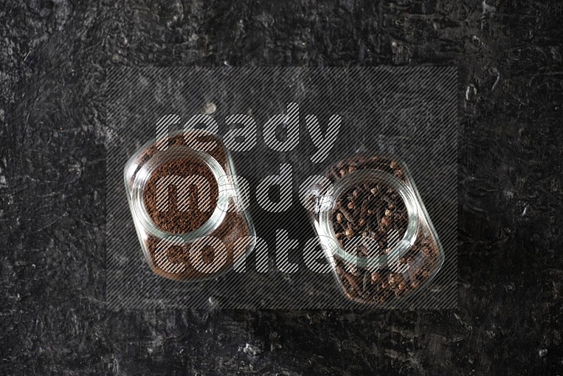 Glass spice jars full of cloves and cloves powder on textured black flooring