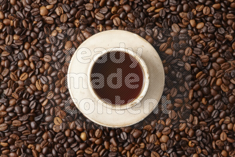 A beige pottery cup of coffee surrounded by roasted coffee beans on beige background