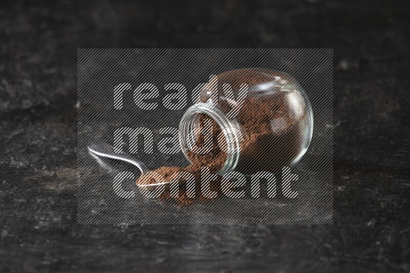 A flipped glass spice jar and a metal spoon full of cloves powder on textured black flooring