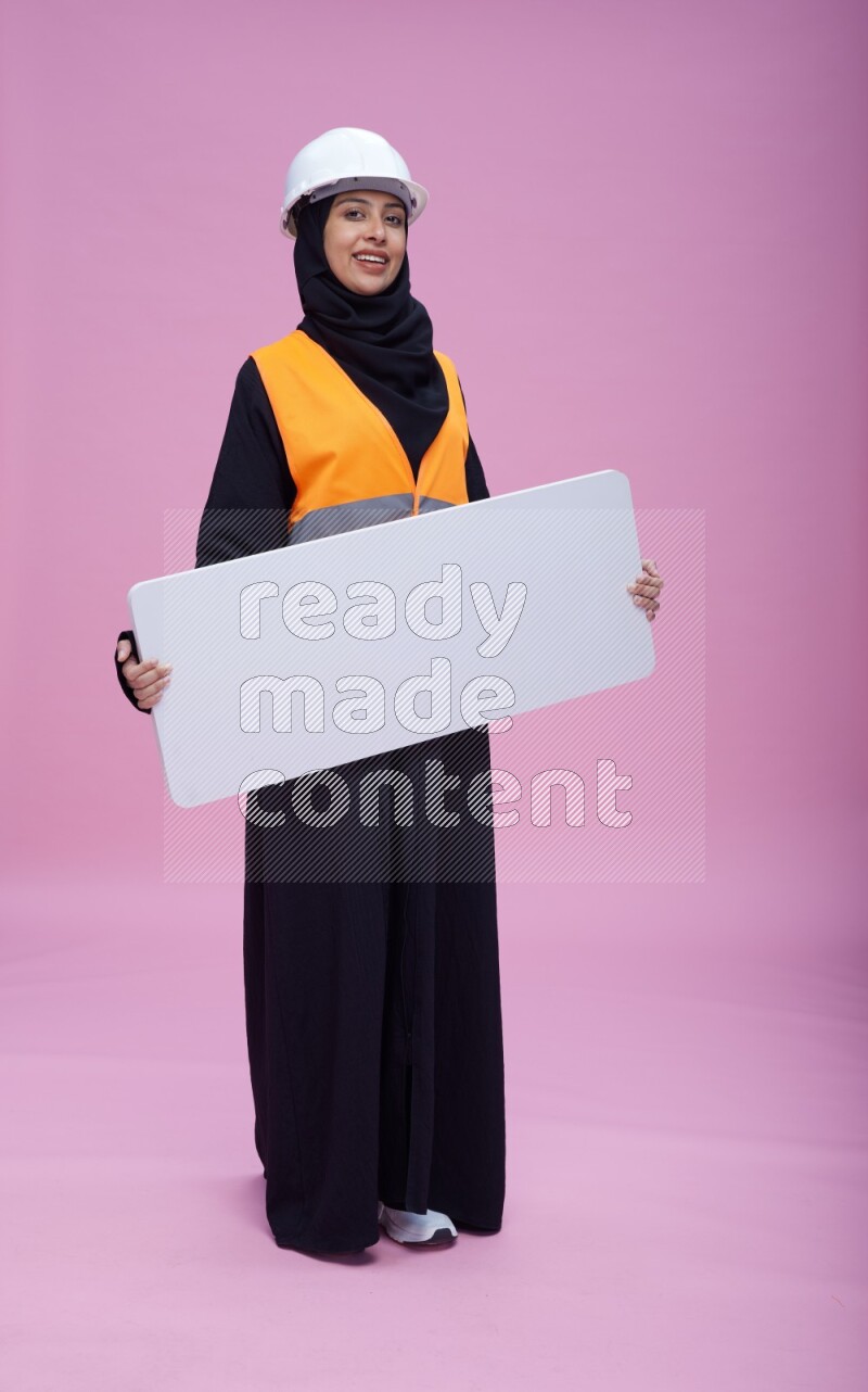 Saudi woman wearing Abaya with engineer vest and helmet standing holding board on pink background