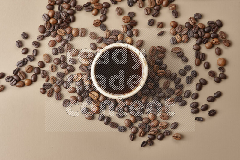 A beige pottery cup of coffee surrounded by roasted coffee beans on beige background