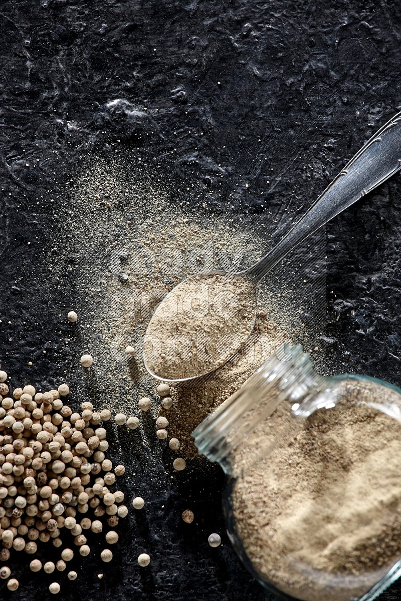 A flipped herbal glass jar and metal spoon full of white pepper powder with spilled powder and pepper beads on textured black flooring