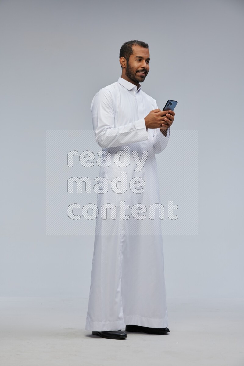 Saudi man wearing Thob standing texting on phone on Gray background