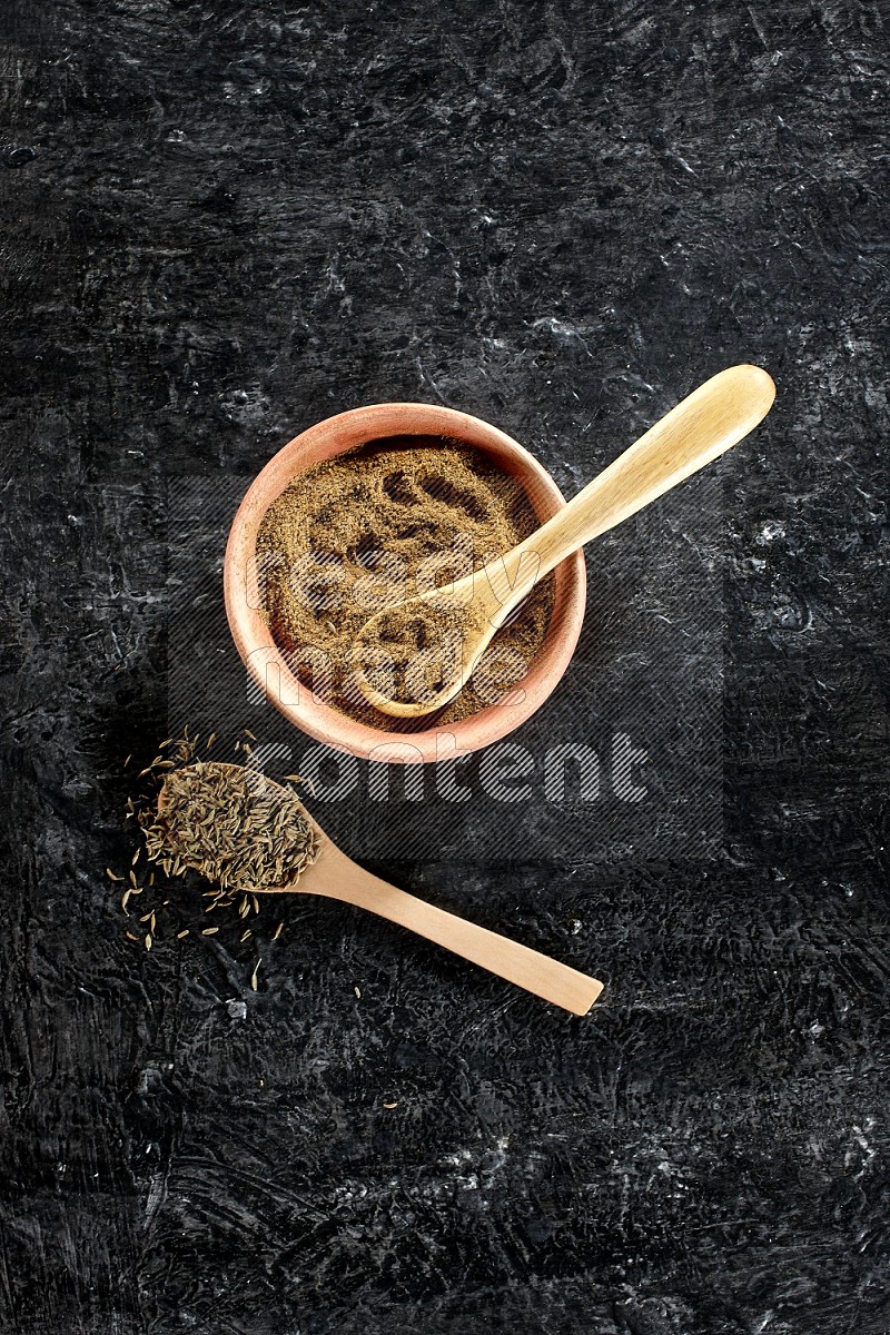 A wooden bowl and 2 wooden spoons full of cumin powder and cumin seeds on a textured black flooring