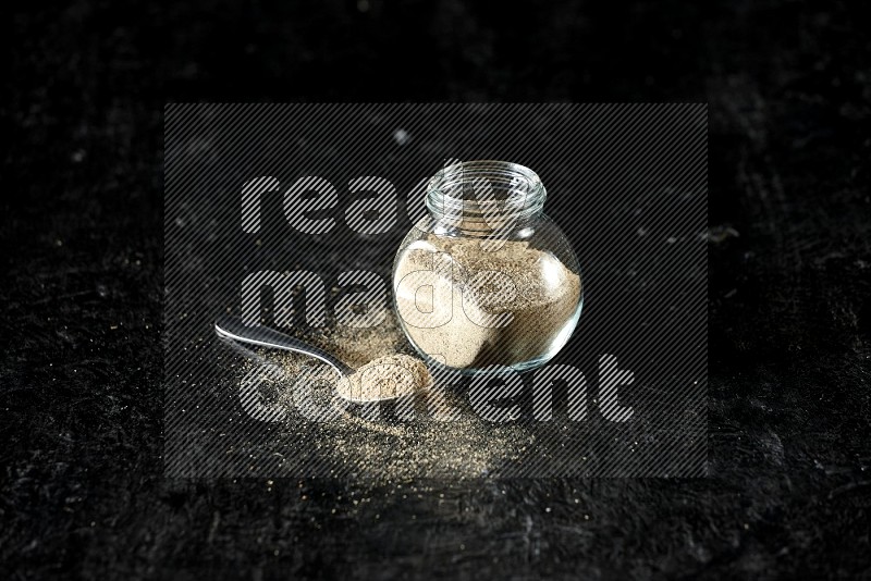 A glass spice jar and metal spoon full of cardamom powder on textured black flooring