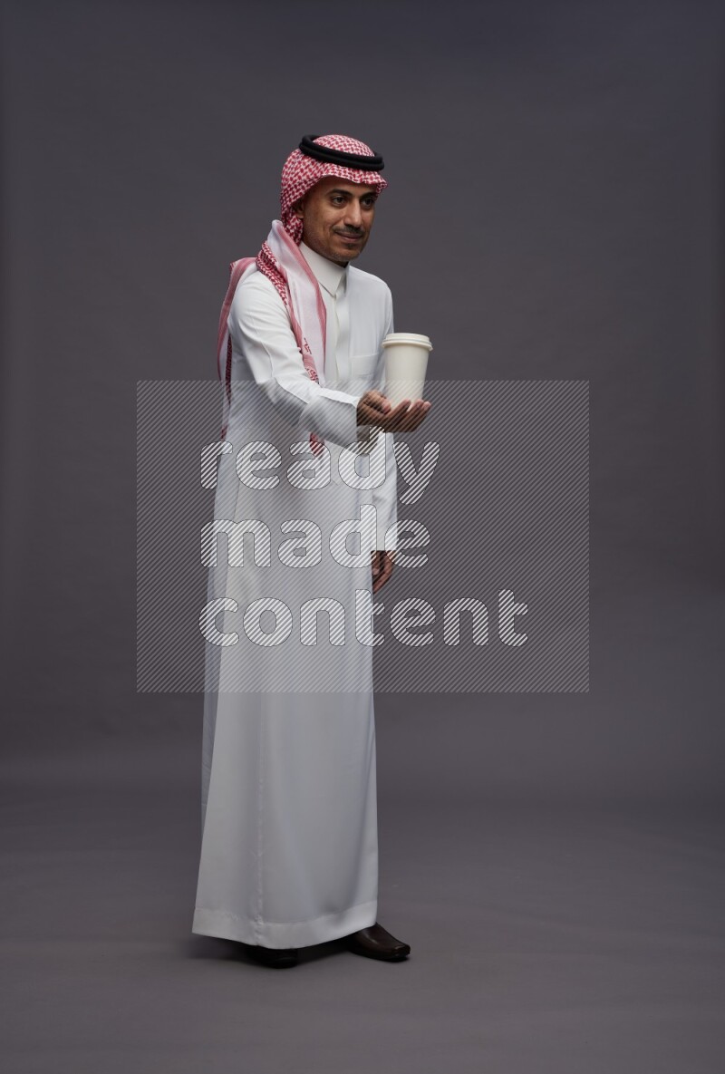 Saudi man wearing thob and shomag standing holding paper cup on gray background