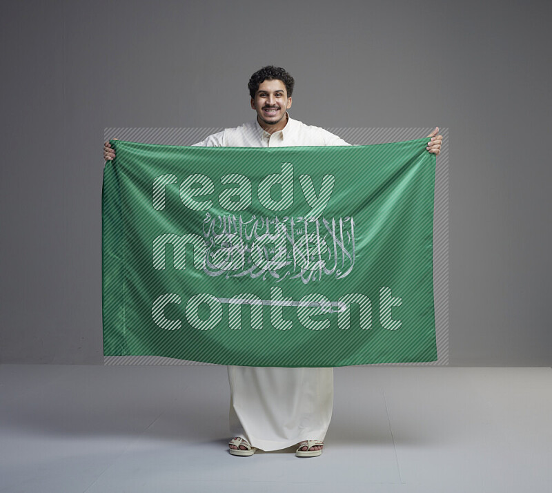 A saudi man standing wearing thob holding big saudi flag on gray background