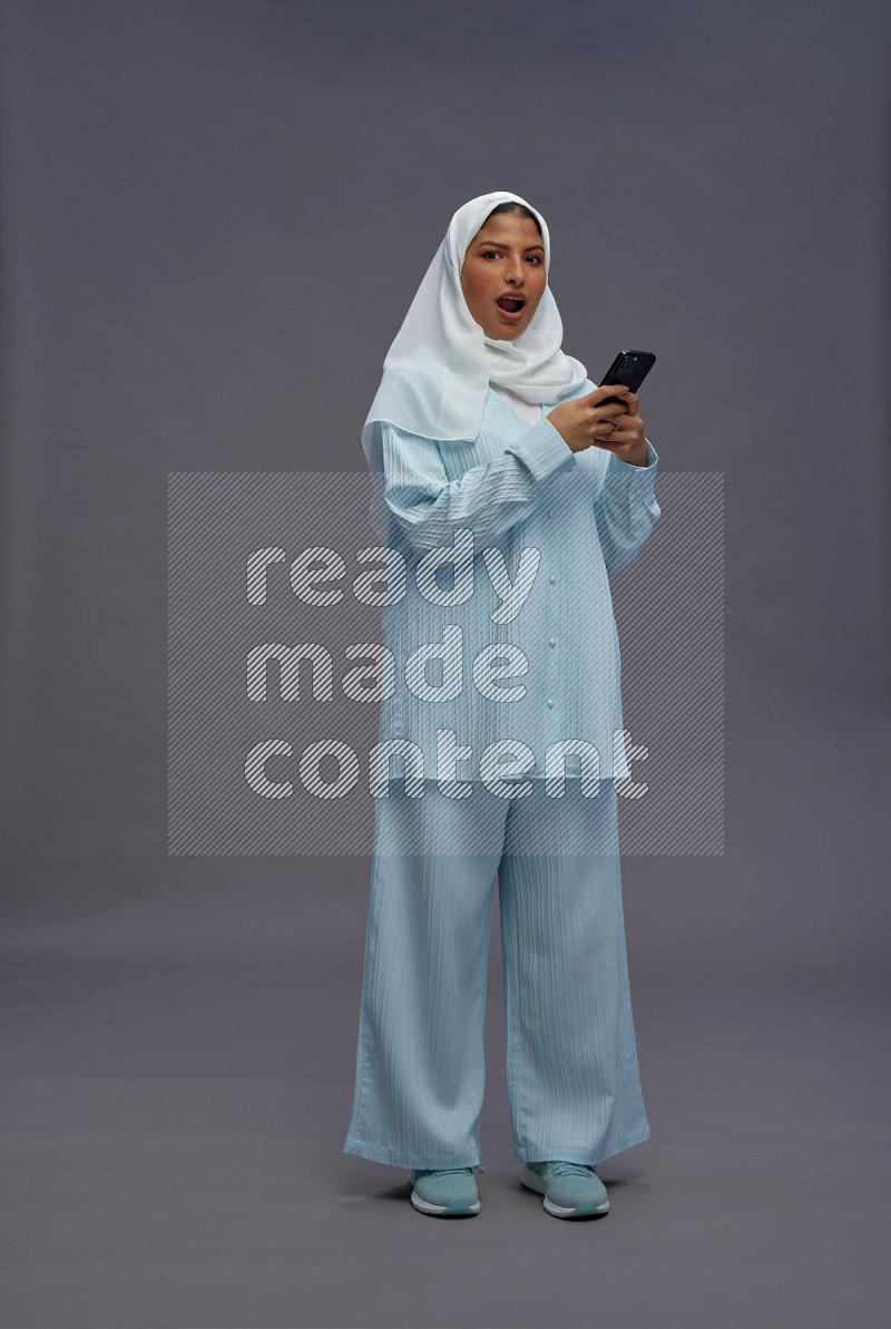 Saudi woman wearing hijab clothes standing texting on phone on gray background