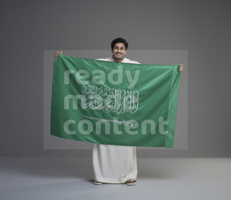 A Saudi man standing wearing thob holding big Saudi flag on gray background
