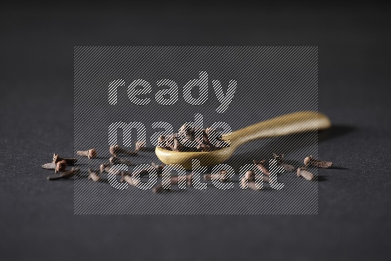 A wooden spoon full of cloves and some spreaded out of it on a black flooring