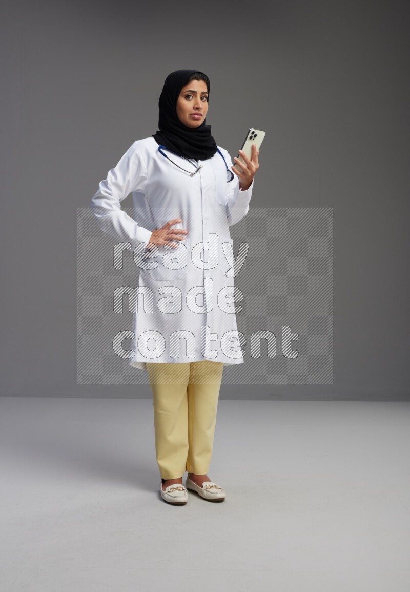 Saudi woman wearing lab coat with stethoscope standing texting on phone on Gray background