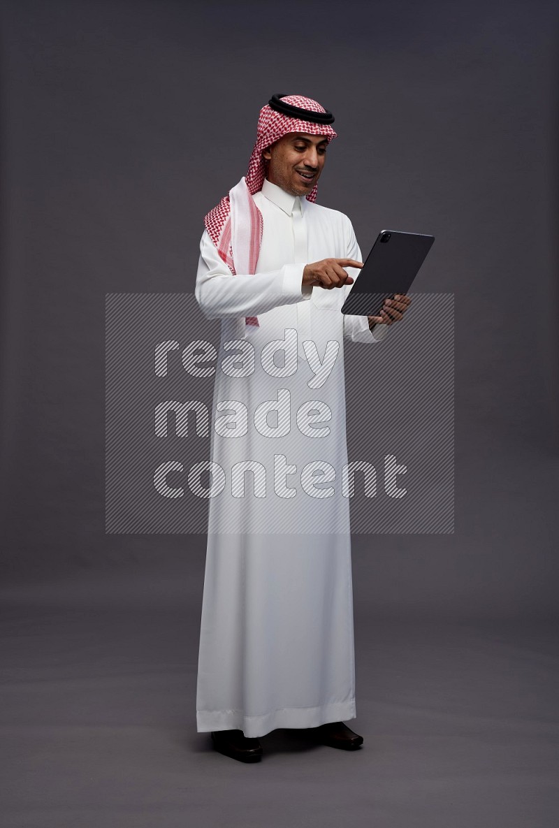 Saudi man wearing thob and shomag standing working on tablet on gray background