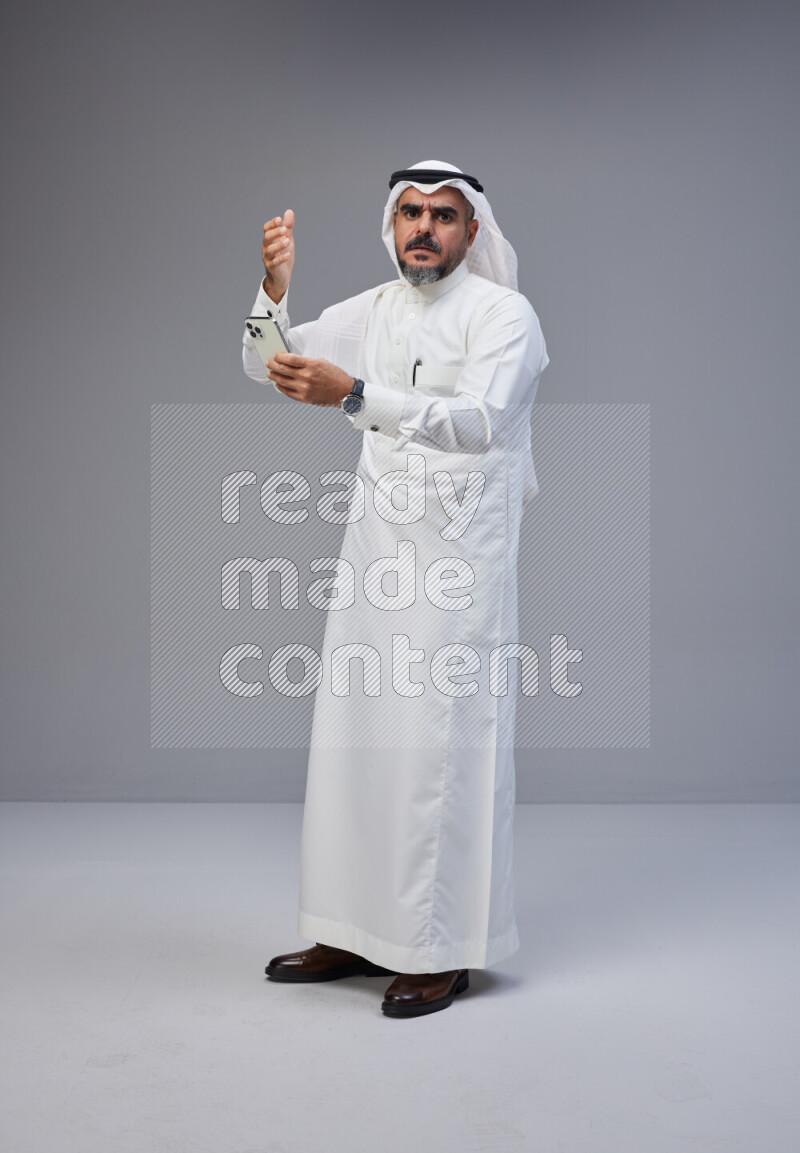 Saudi man Wearing Thob and white Shomag standing texting on phone on Gray background