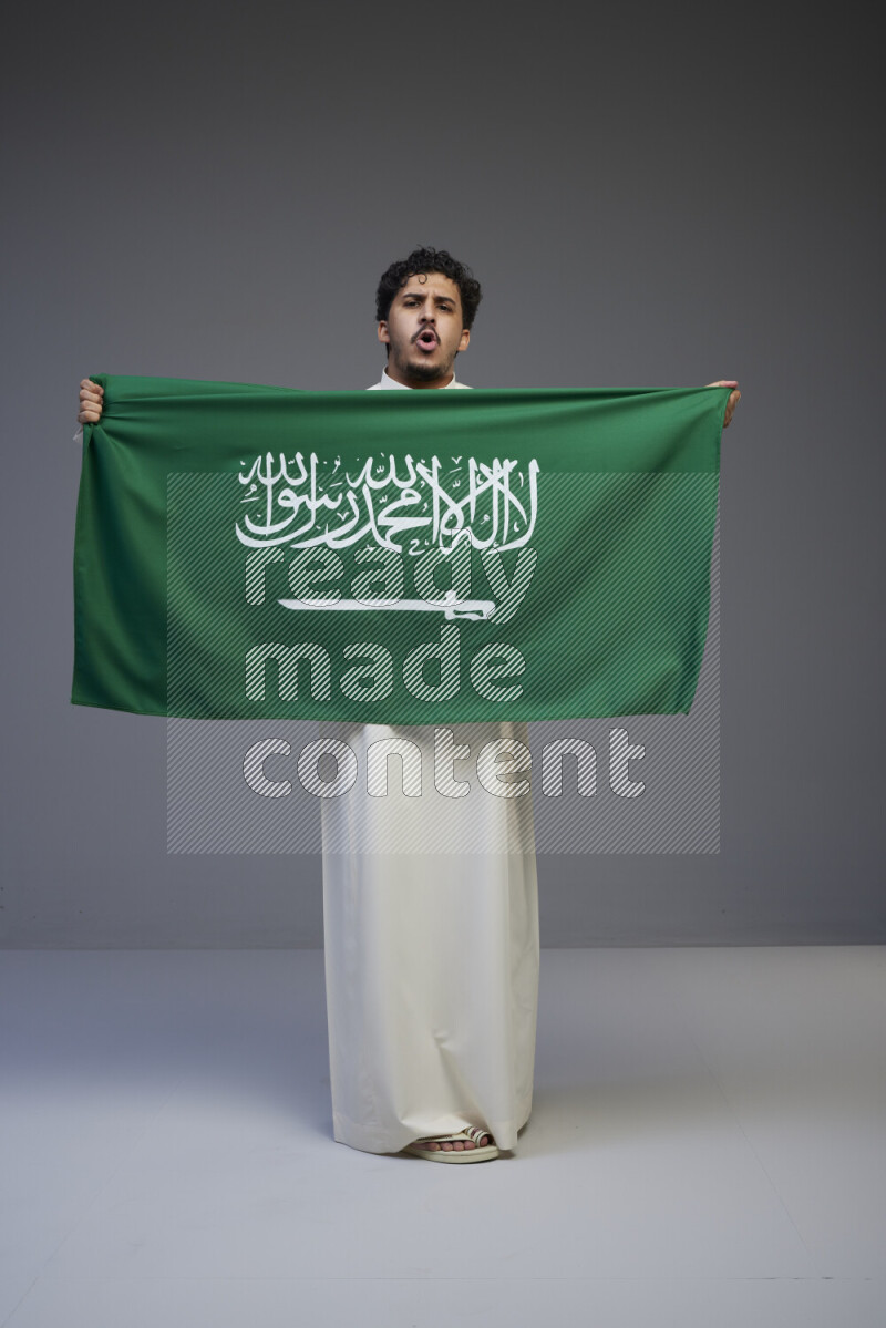 A Saudi man standing wearing thob holding big Saudi flag on gray background
