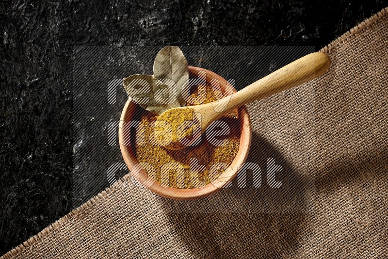A wooden bowl and a wooden spoon full of turmeric powder on burlap fabric on textured black flooring