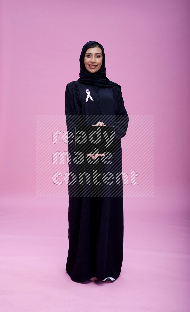 Saudi woman wearing pink ribbon on Abaya standing showing tablet to camera on pink background