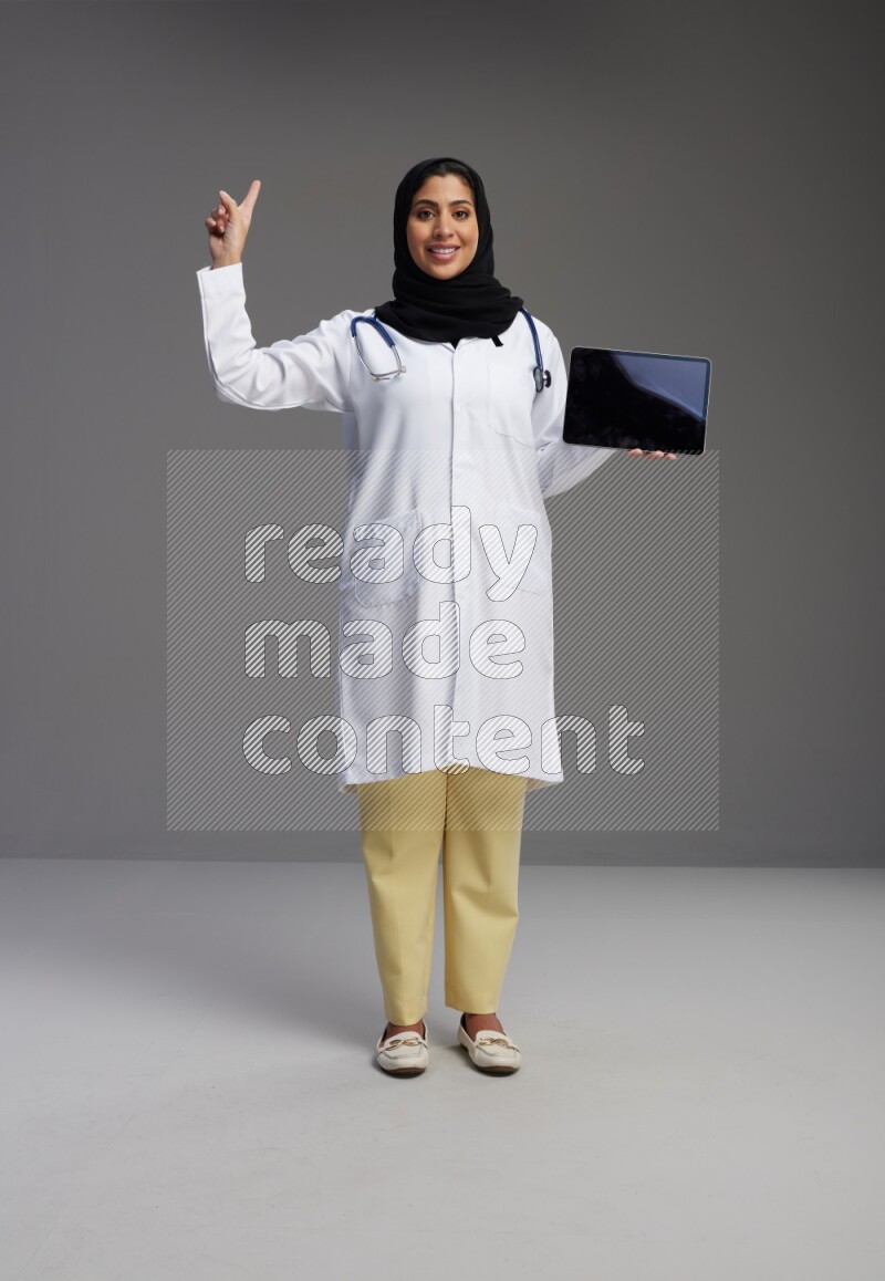 Saudi woman wearing lab coat with stethoscope standing showing tablet to camera with sign in the back on Gray background