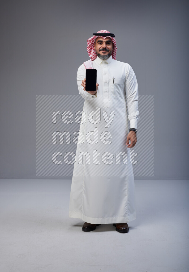 Saudi man Wearing Thob and red Shomag standing showing phone to camera on Gray background