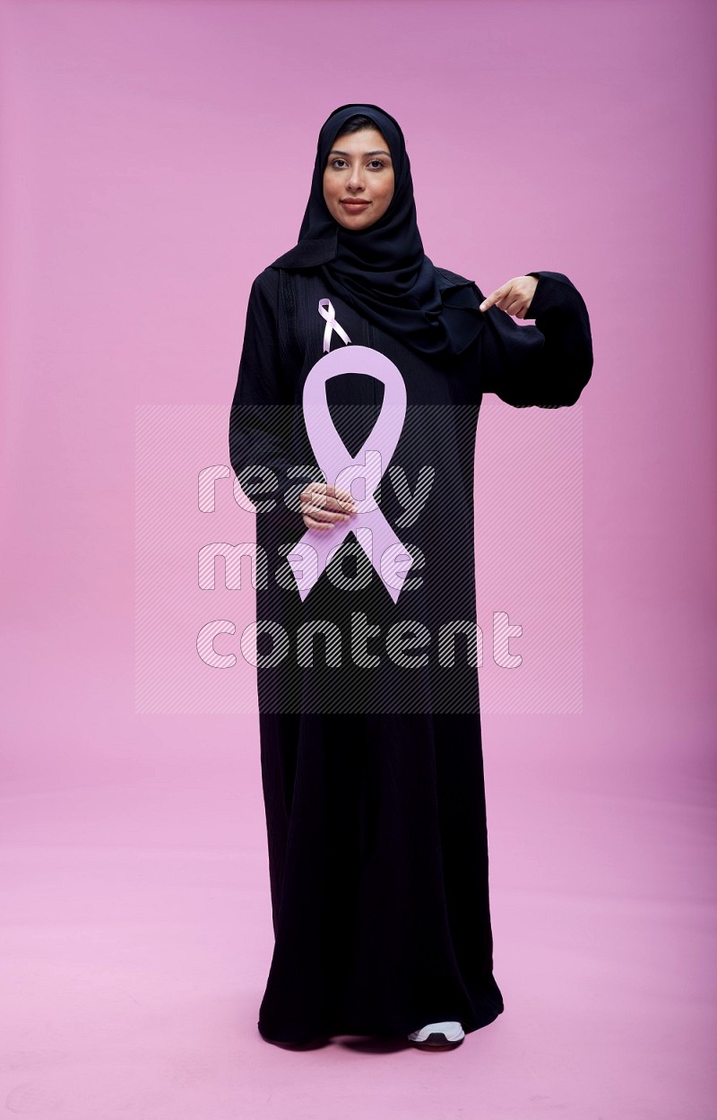 Saudi woman wearing pink ribbon on abaya standing holding awareness ribbon on plain pink background