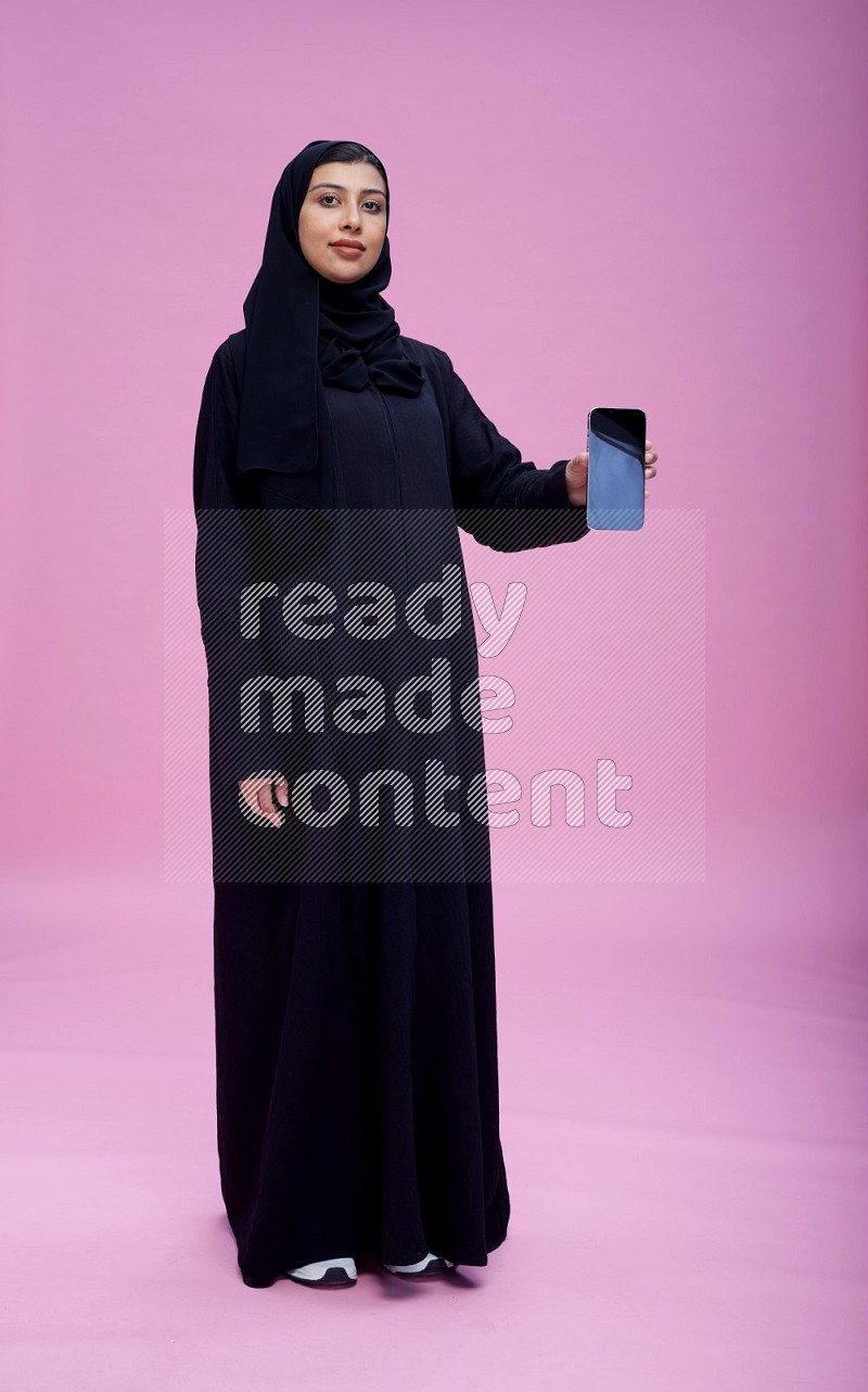 Saudi woman wearing Abaya standing showing phone to camera on pink background