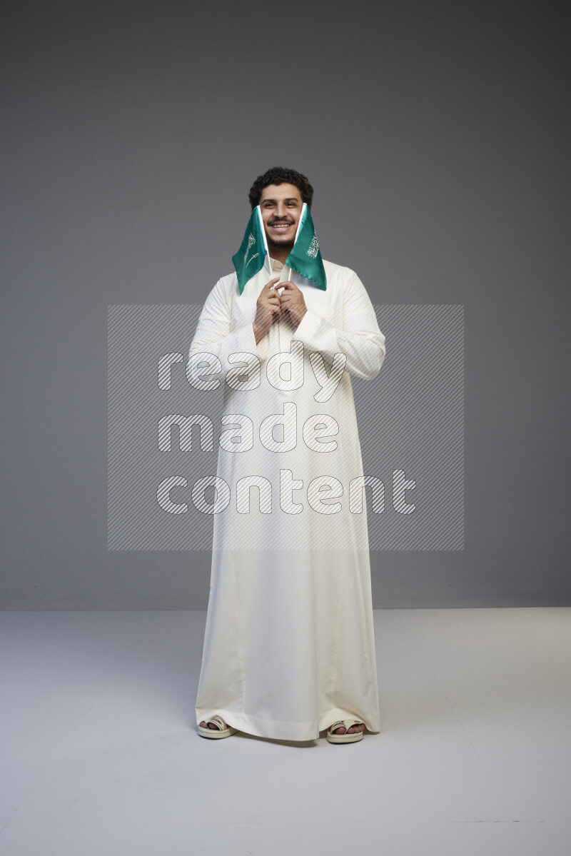 A Saudi man standing wearing thob holding small Saudi flag on gray background