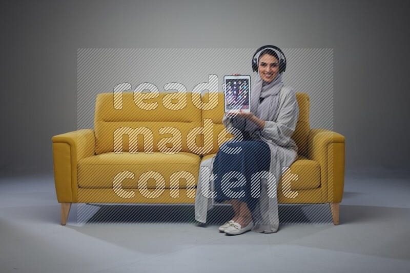 A Saudi female wearing a light gray Abaya and head scarf sitting on a yellow sofa and showing her tablet's screen while wearing headphones eye level on a grey background
