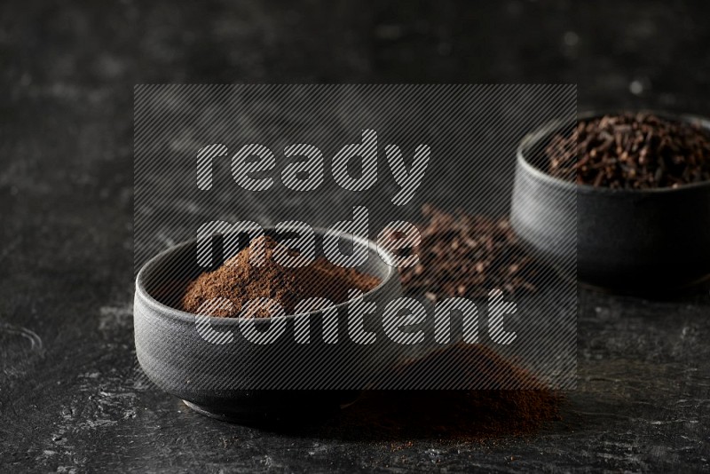 2 Black pottery bowls full of cloves and the other full of cloves powder on textured black flooring