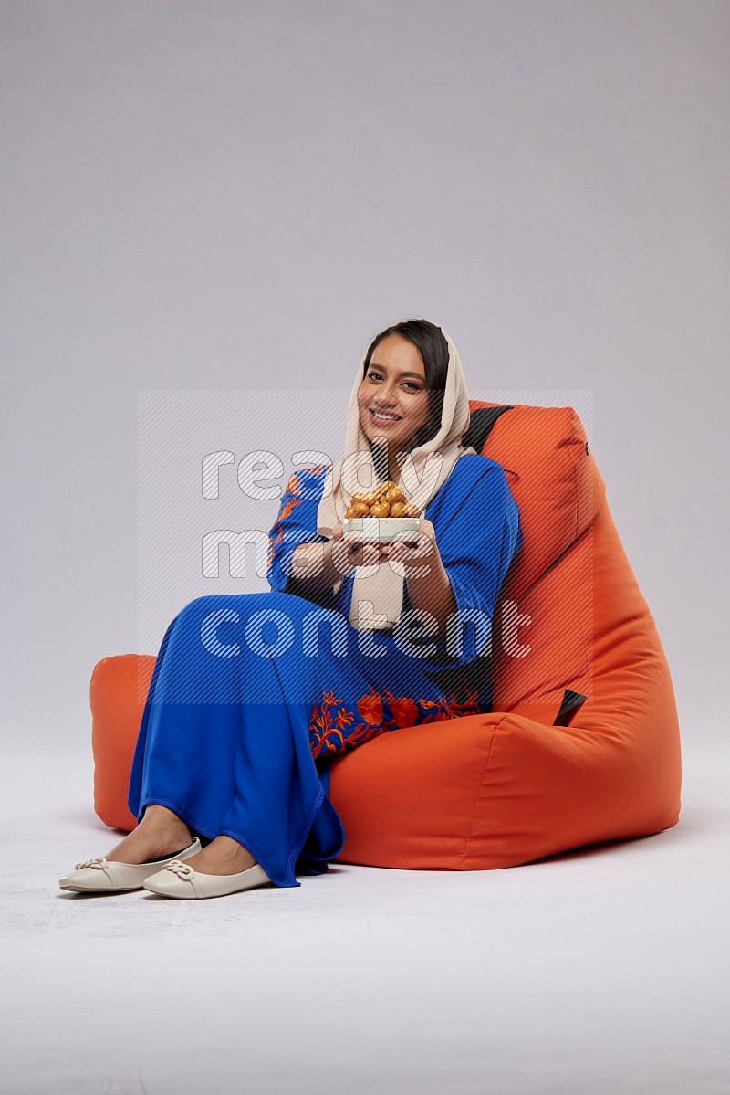 A Woman sitting on an orange beanbag wearing Jalabeya holding a plate of luqaimat