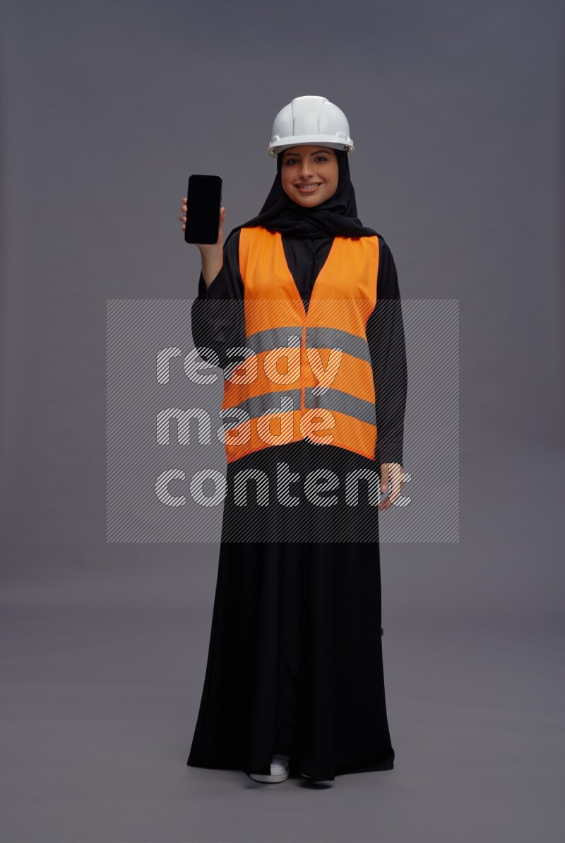Saudi woman wearing Abaya with engineer vest standing showing phone to camera on gray background