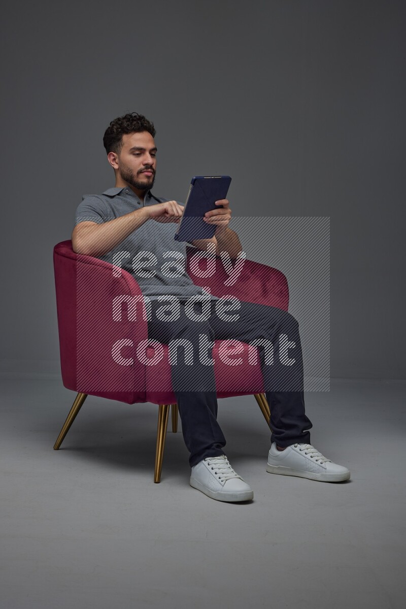 A man wearing casual and using his tablet while sitting on a burgundy chair eye level on a gray background