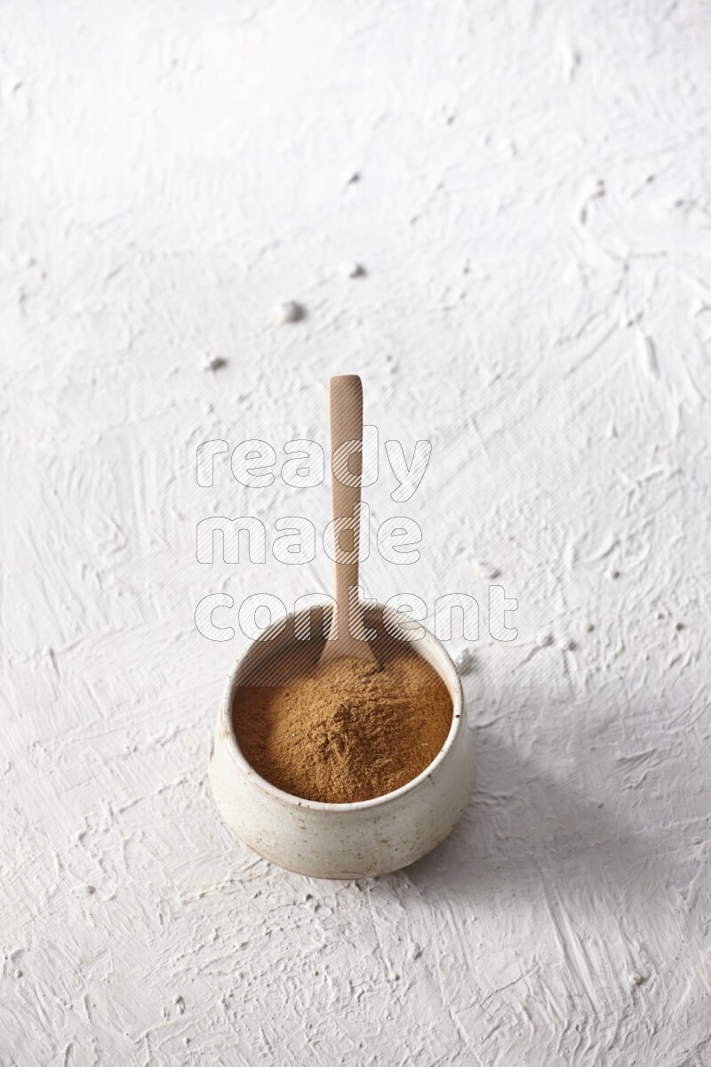 Ceramic beige bowl full of cinnamon powder with a wooden spoon on a textured white background