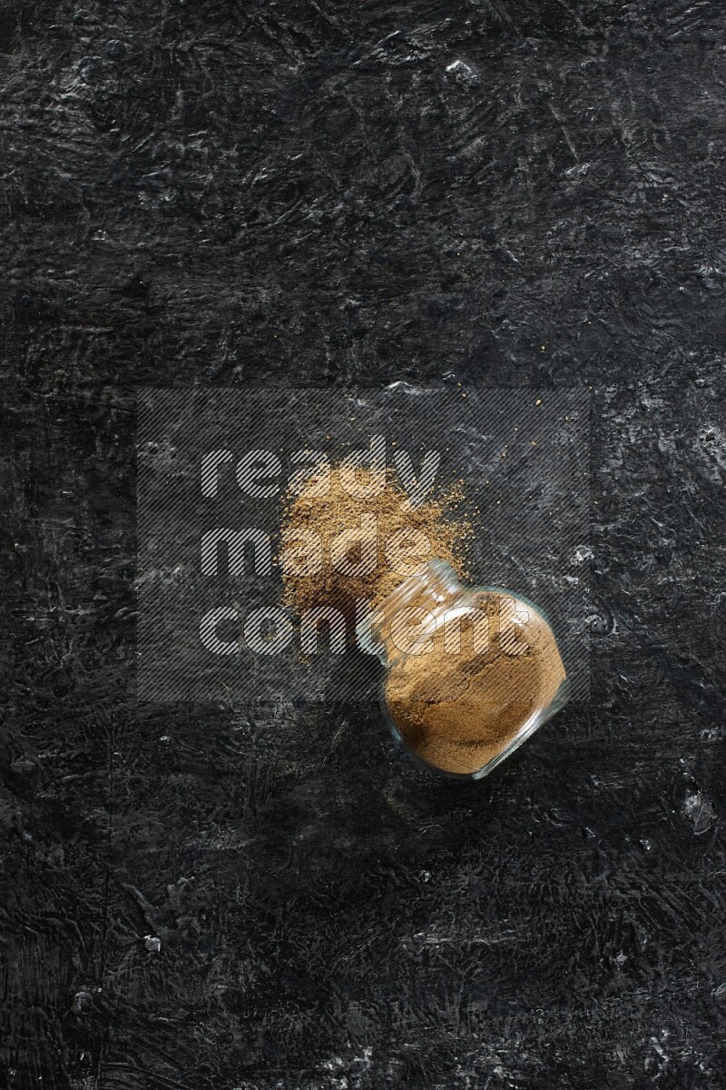 A flipped glass spice jar full of cumin powder and powder spilled out on a textured black flooring