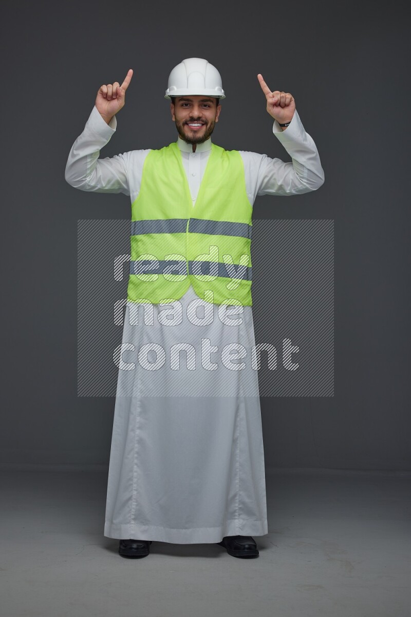 A Saudi man wearing Thobe with a yellow safety vest and white helmet standing and pointing different angles eye level on a gray background