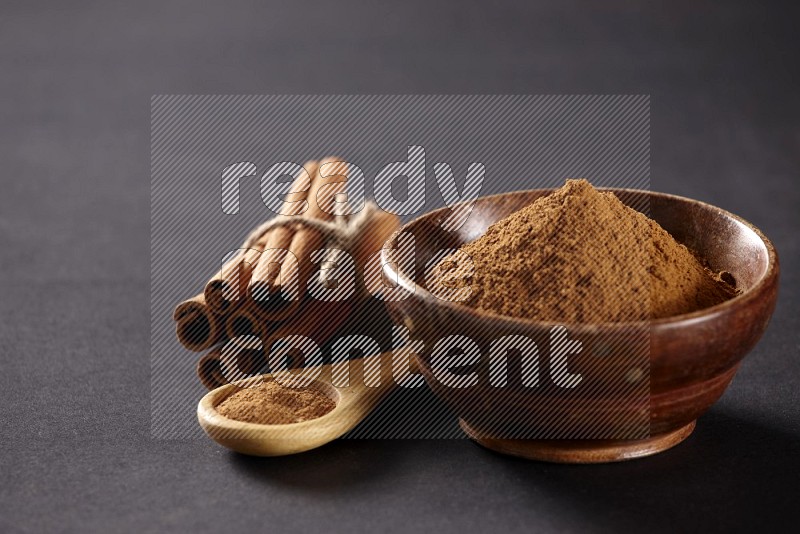 Cinnamon sticks stacked and bounded beside a wooden bowl full of cinnamon powder and a wooden spoon full of powder on black background
