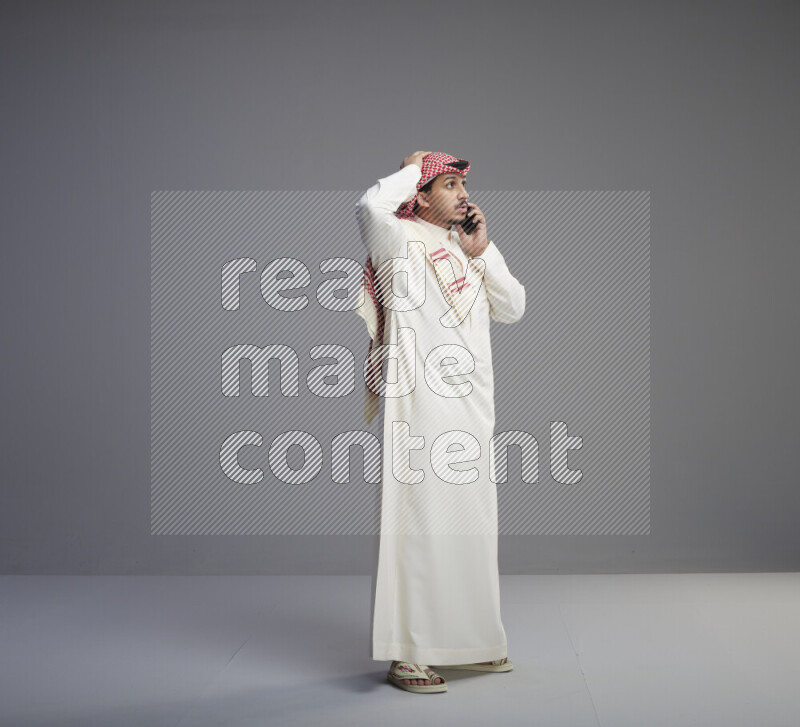 A Saudi man standing wearing thob and red shomag talking on phone on gray background
