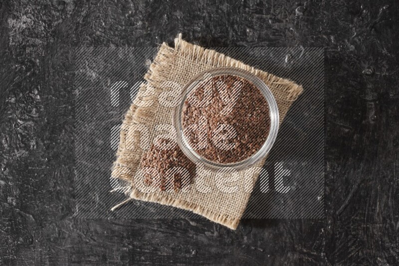 A glass bowl full of flaxseeds with bunch of the seeds on burlap fabric on a textured black flooring