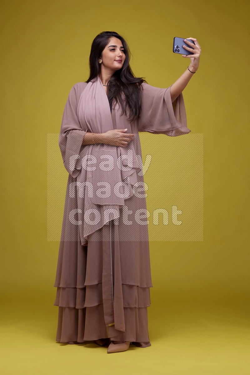 A woman Taking a Selfie on a Yellow Background wearing Brown Abaya