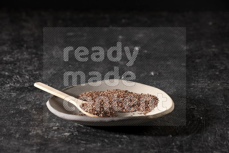 A multicolored pottery plate full of flaxseeds and wooden spoon full of seeds on a textured black flooring