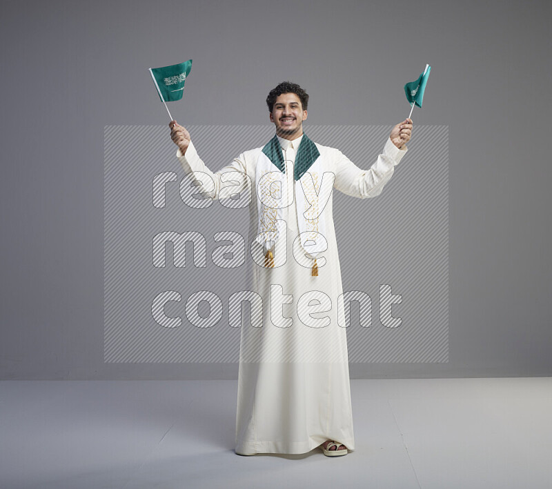 A Saudi man standing wearing thob and saudi flag scarf and holding small saudi flag on gray background