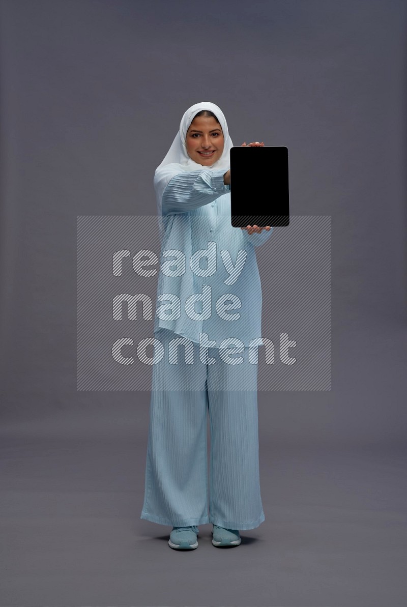 Saudi woman wearing hijab clothes standing showing tablet to camera on gray background