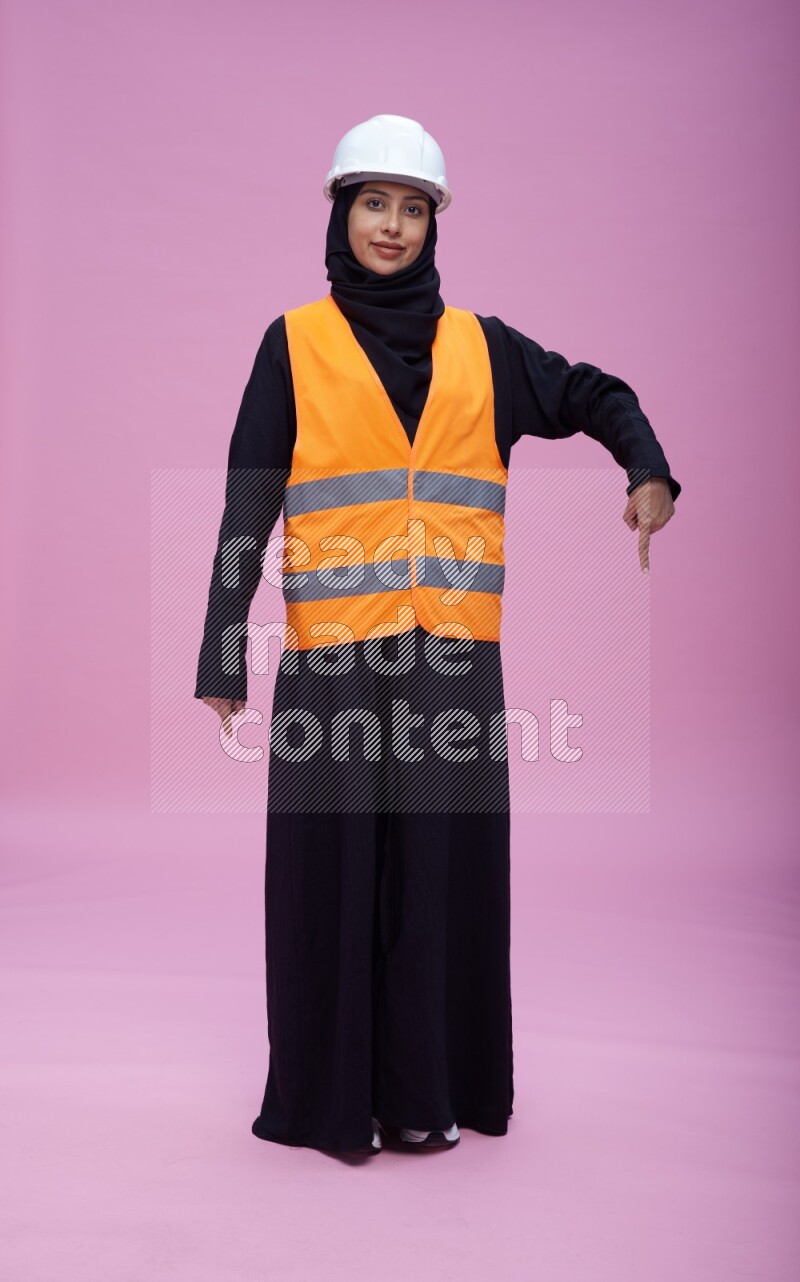 Saudi woman wearing Abaya with engineer vest and helmet standing interacting with the camera on pink background
