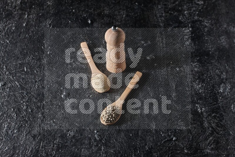 2 wooden spoons one full of white pepper powder and the other with pepper beads and a wooden pepper mill on textured black flooring