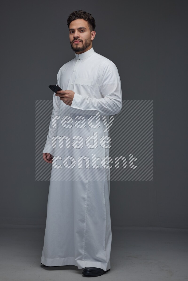 A Saudi man wearing Thobe standing and using his phone eye level on a gray background