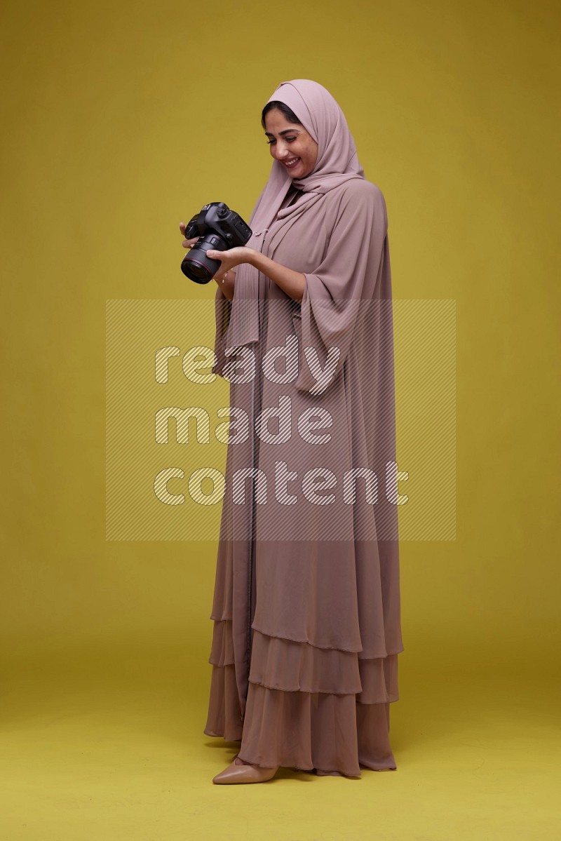 A woman Holding a Camera on a Yellow Background wearing Brown Abaya with Hijab