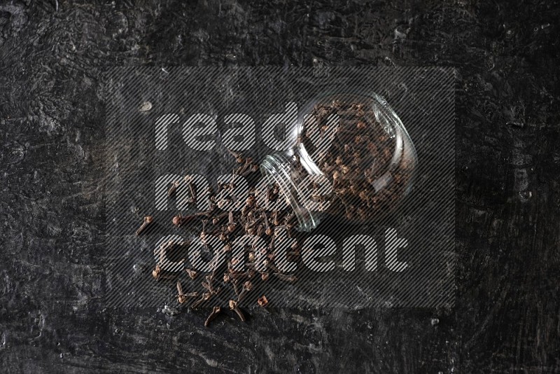 A flipped glass spice jar full of cloves and cloves came out of it on textured black flooring