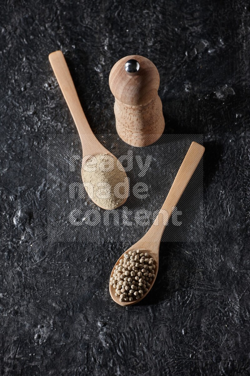 2 wooden spoons one full of white pepper powder and the other with pepper beads and a wooden pepper mill on textured black flooring