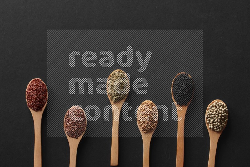 wooden spoons filled with white peppers, mustard seeds, black seeds, garden cress, cumin and flax on black flooring