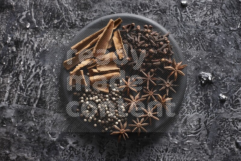 Cinnamon sticks, cloves, star anise and black and white peppers on a black plate on textured black background